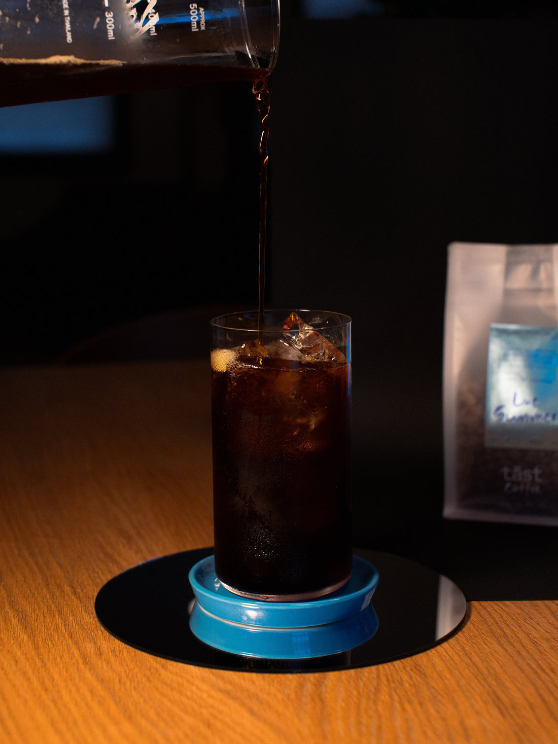 Tall drinking glass full of ice and coffee while coffee is being poured from a carafe above