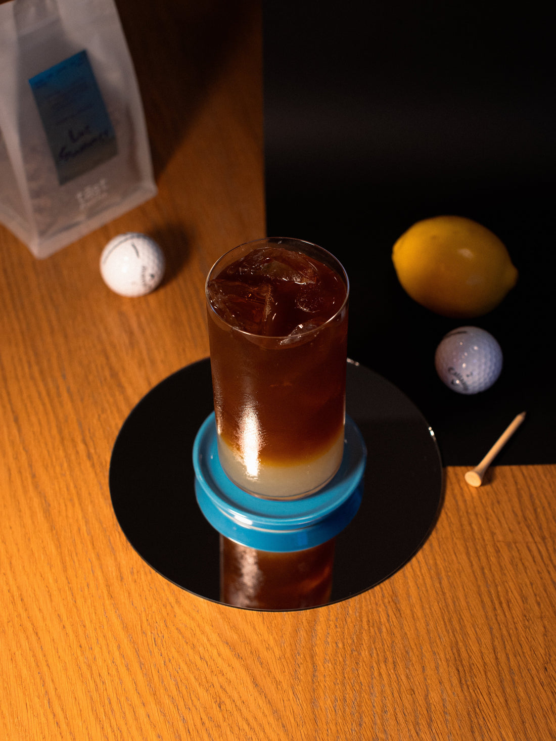 Tall glass with lemonade on the bottom and coffee on the top. Golf balls and lemons scattered on the table around the glass
