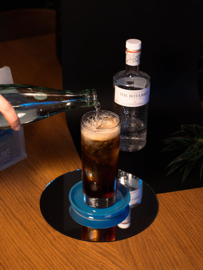 Tall glass filled with fizzing brown liquid with a had pouring sparkling water into the glass
