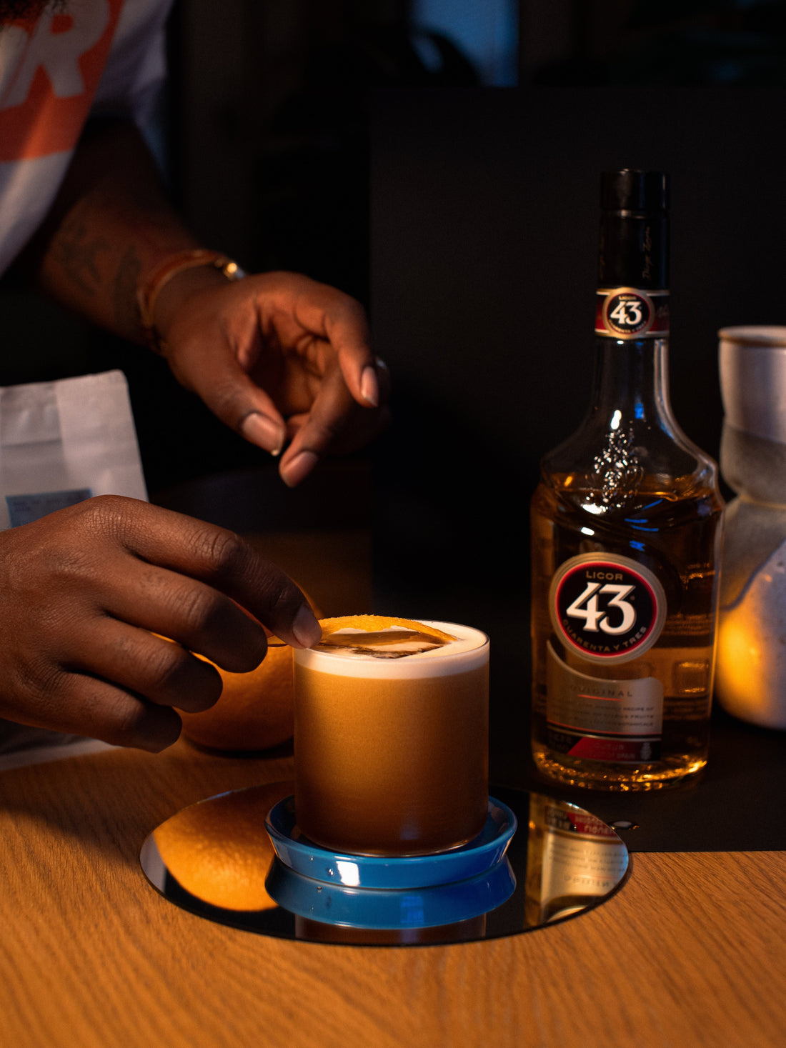 two hands place an orange peel on a short, stemless cocktail glass with a brown drink with foam