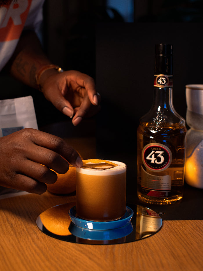 two hands place an orange peel on a short, stemless cocktail glass with a brown drink with foam