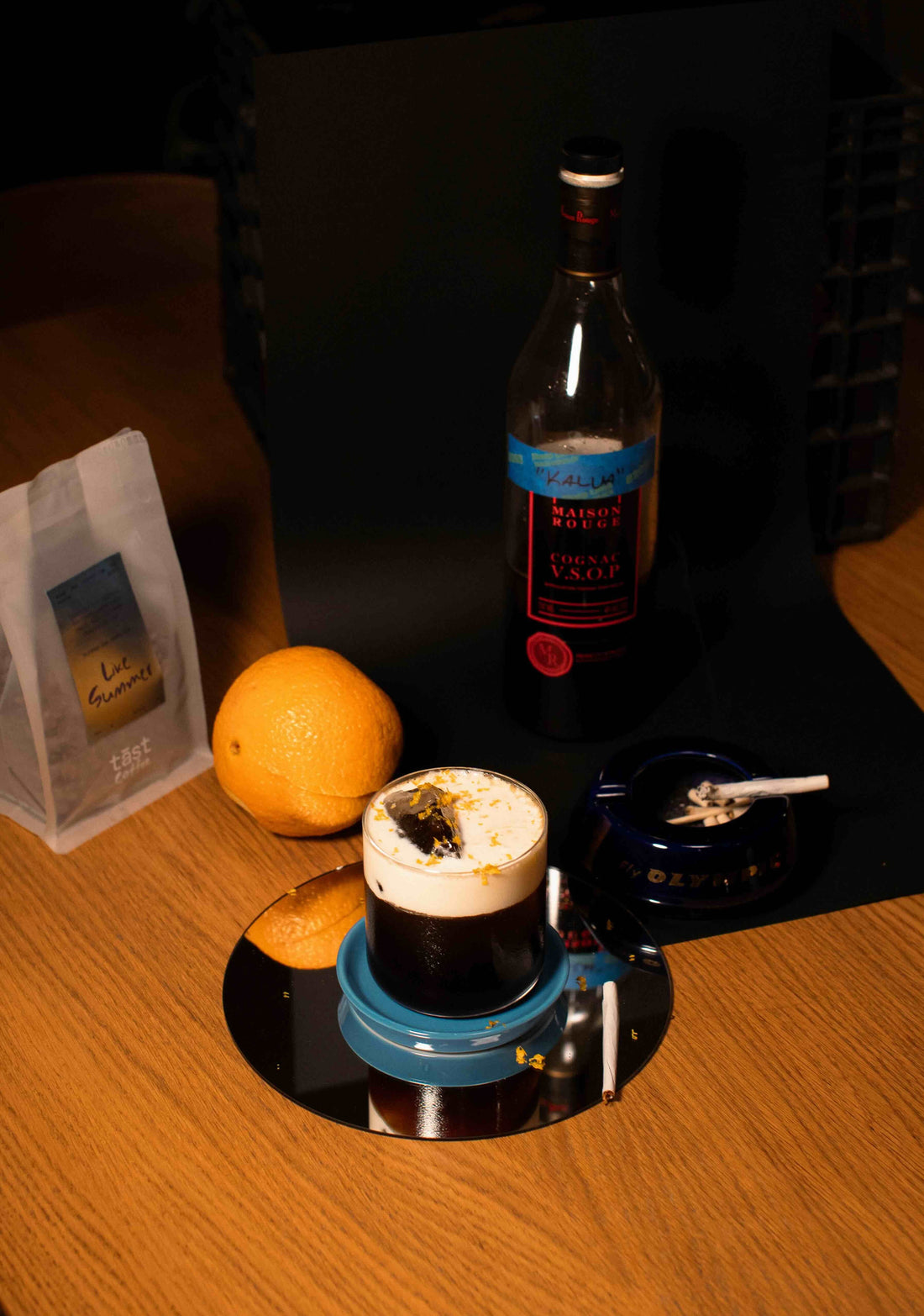 dark brown cocktail in a stemless glass with a large head of foam. An orange sits on the table next to it.
